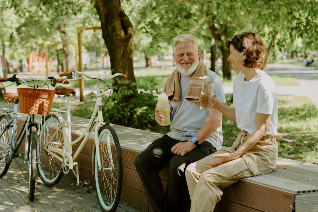 Senior couple resting on wooden bench and drinking water after riding bikes around park